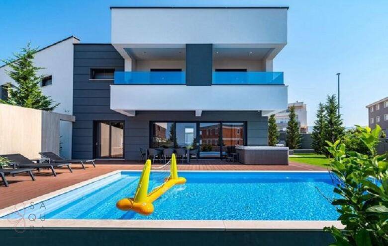 Moderne Villa mit großem Pool, Holzterrasse und aufblasbarem Spielzeug im Gartenbereich.