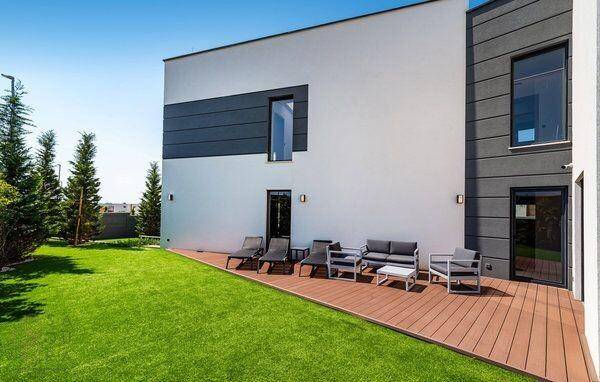 Moderne Hausfassade mit Holzterrasse, Rasenfläche und bequemen Outdoor-Sitzmöbeln unter klarem Himmel.