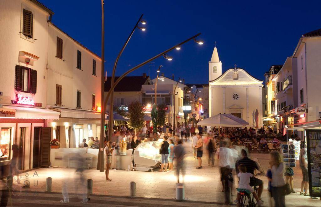 Beleuchteter Stadtplatz bei Nacht mit vielen Menschen, Restaurants und einer Kirche im Hintergrund.