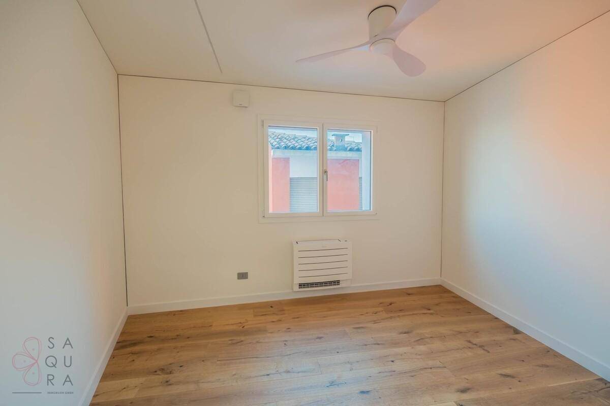 Helles Schlafzimmer mit Holzboden, Deckenventilator und einem Fenster mit Blick auf die Umgebung.