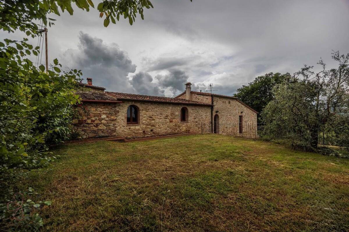 Historisches Steinhaus umgeben von Bäumen und üppiger Vegetation unter bewölktem Himmel.