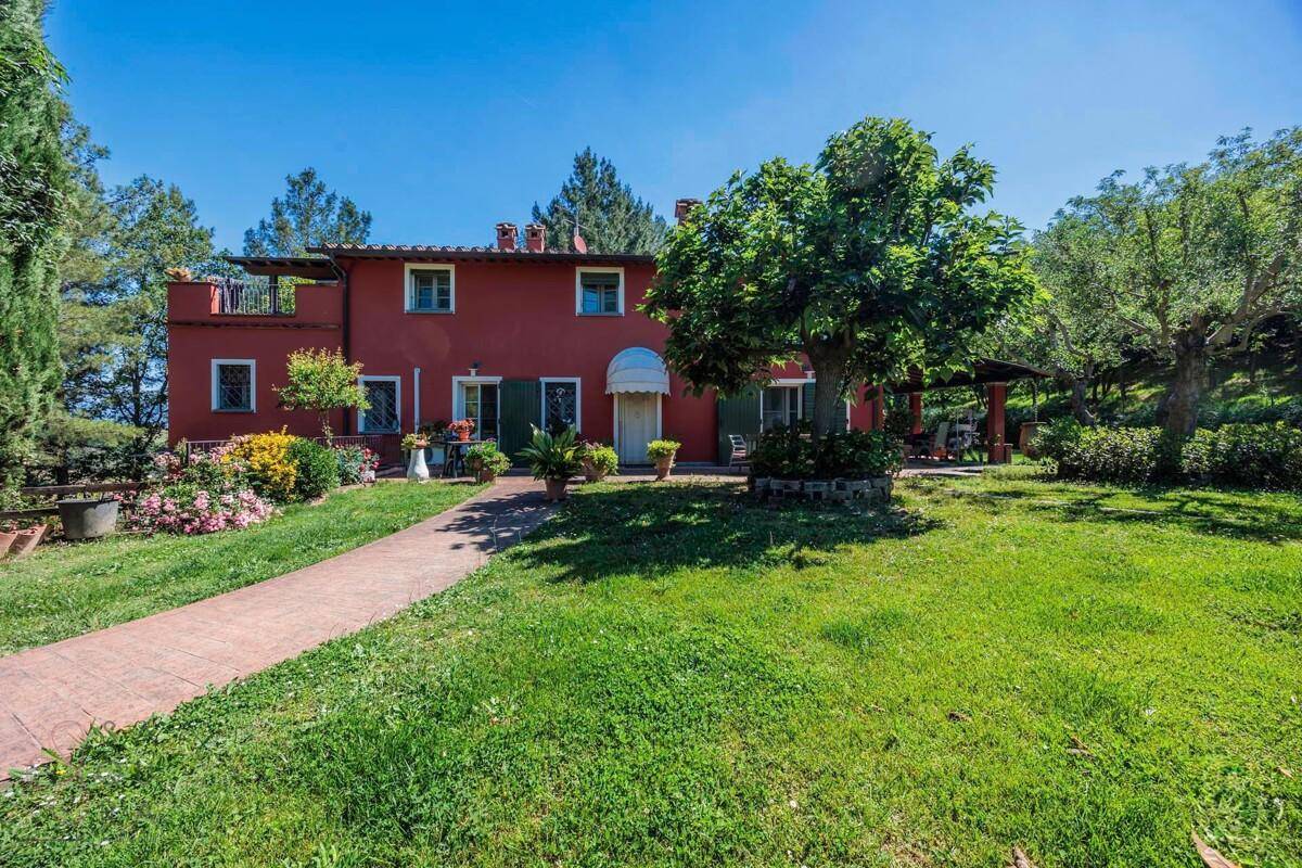 Weitläufiger Blick auf die Vorderseite des roten Hauses mit Rasenfläche, gepflastertem Weg und überdachtem Sitzbereich.