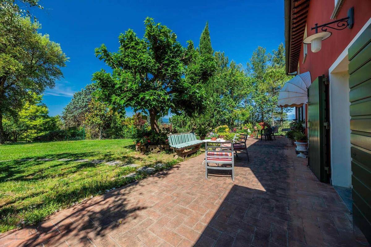 Gepflasterte Terrasse mit Gartenmöbeln neben dem roten Haus, umgeben von üppigem Grün.