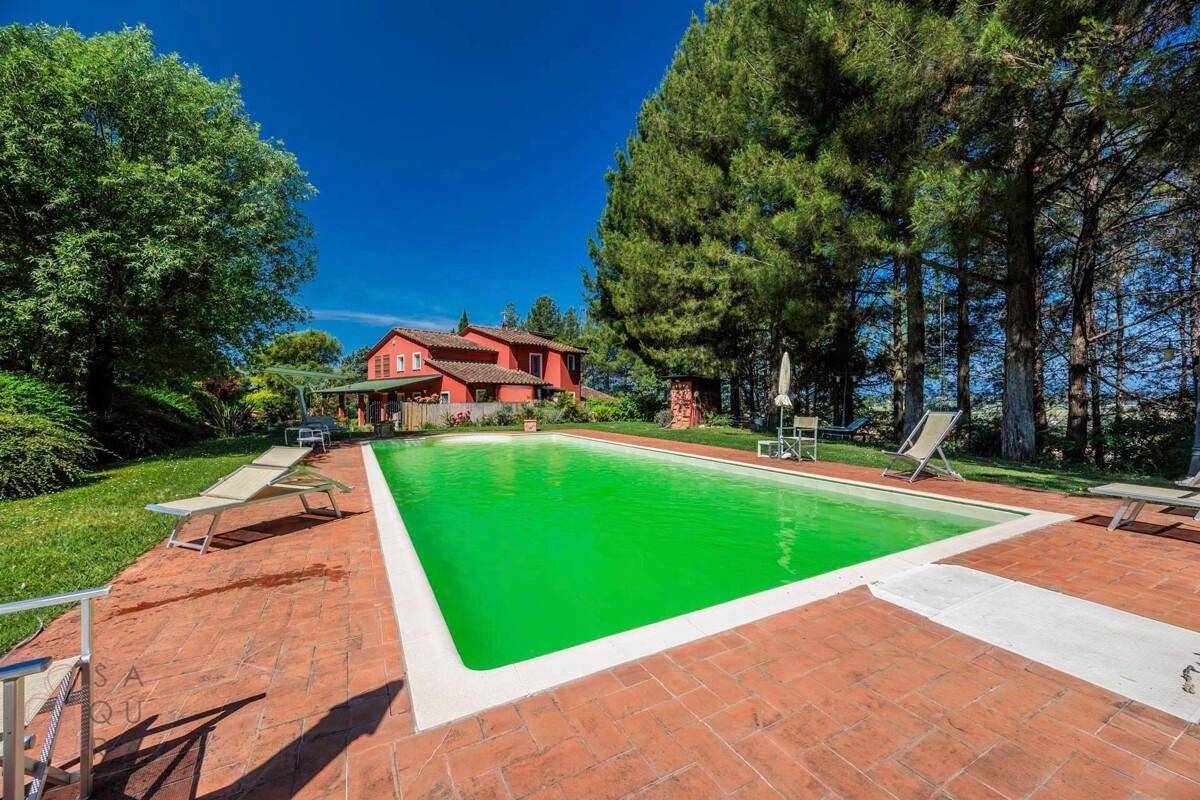 Großer Swimmingpool mit Terrakotta-Pflasterung und Liegestühlen, das rote Haus im Hintergrund.