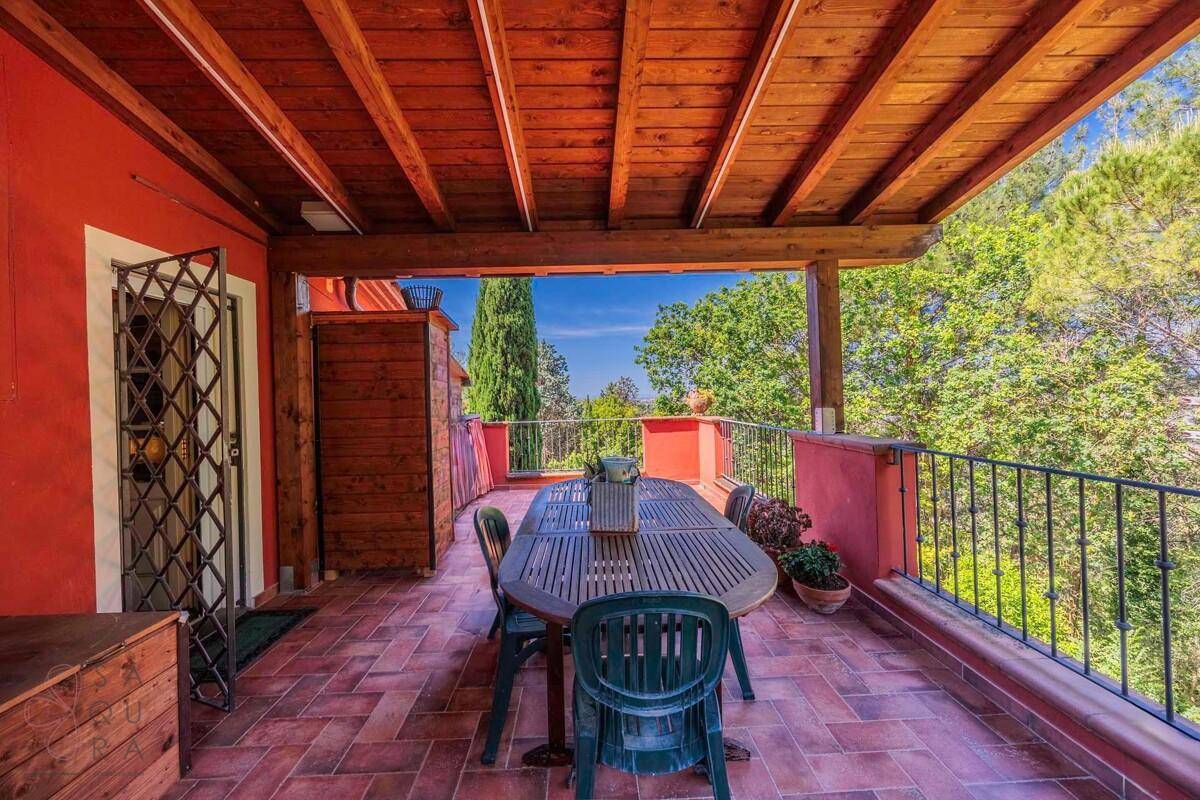 Geräumige überdachte Terrasse mit Holztisch und Stühlen, umgeben von üppiger Vegetation und Fernblick.