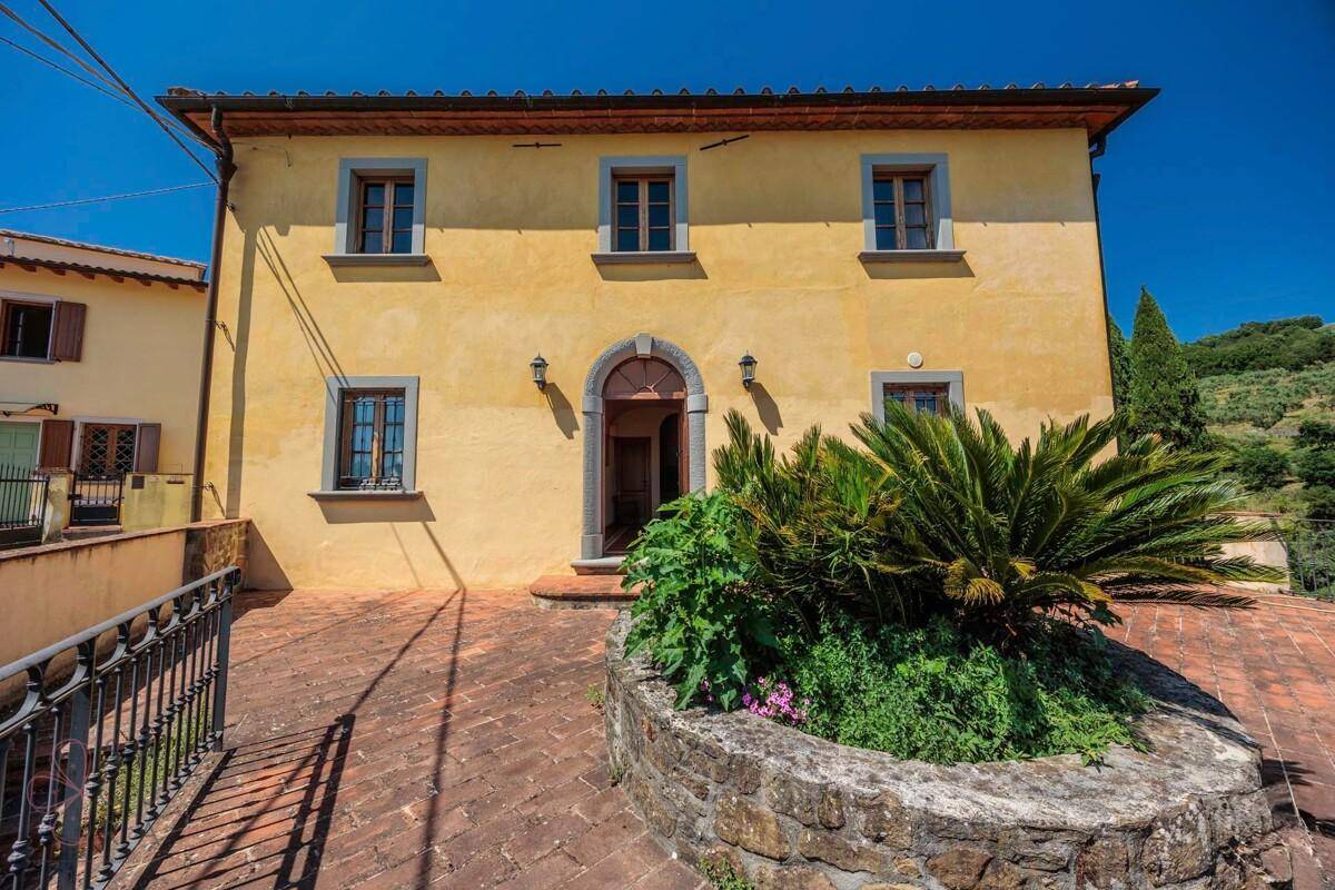 Gelbe Fassade des traditionellen Hauses mit gepflastertem Vorplatz und mediterraner Bepflanzung.