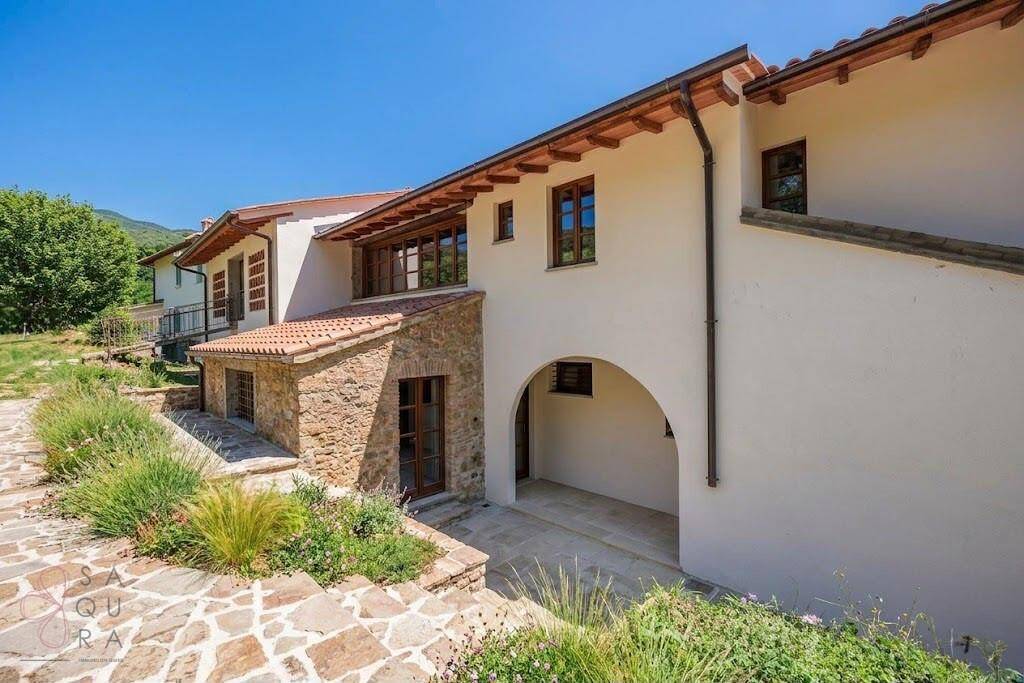 Seitenansicht des Hauses mit Natursteinfassade, gepflastertem Weg und mediterraner Bepflanzung.