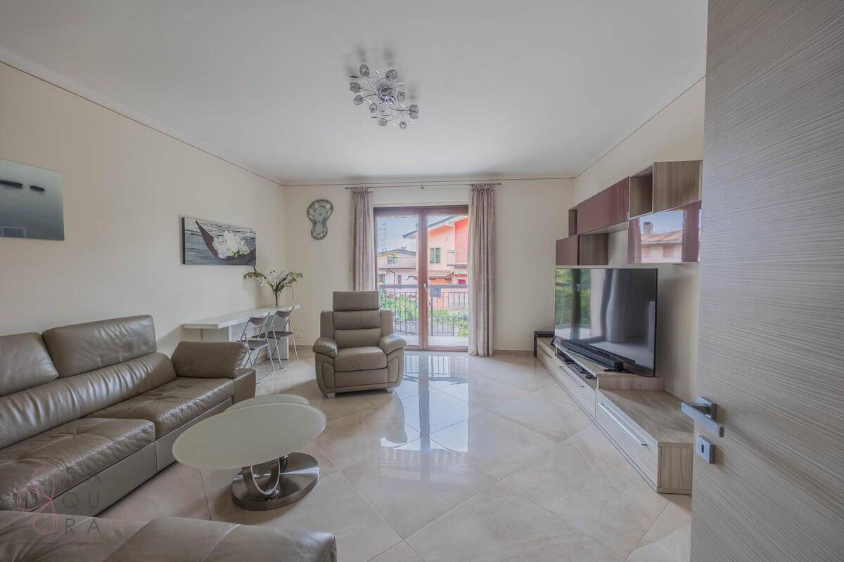 Helles Wohnzimmer mit beigem Ledersofa, Fliesenboden und Zugang zum Balkon.