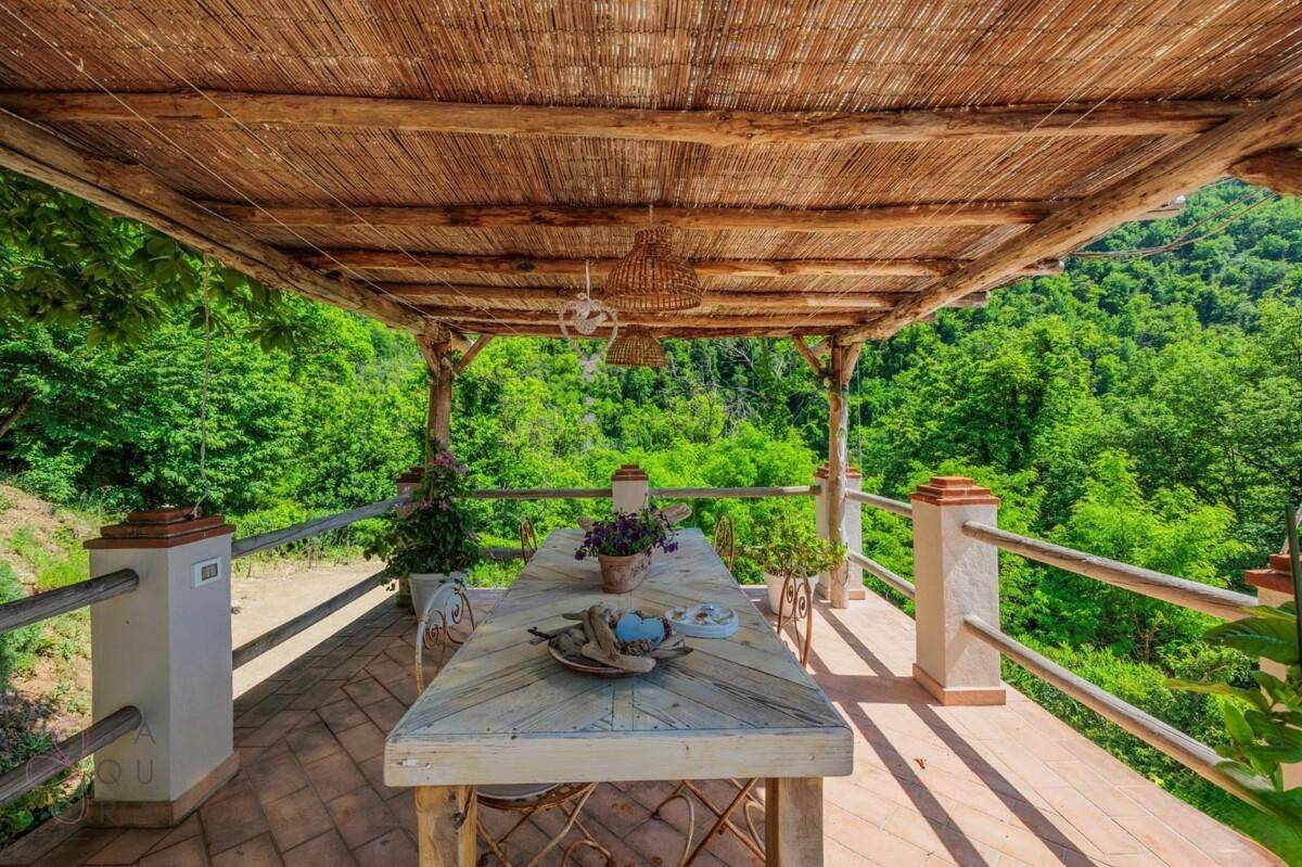 Geräumige überdachte Terrasse mit Holztisch und Stühlen, umgeben von üppiger Natur und Bergblick.
