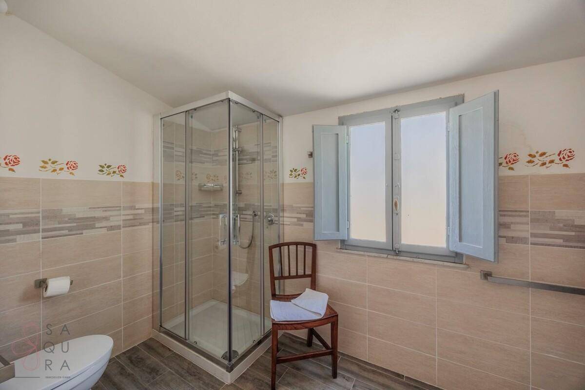 Helles Badezimmer mit Duschkabine, beigen Fliesen und Fenster mit Fensterläden.