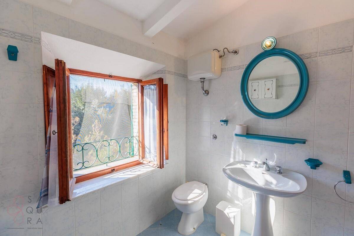 Badezimmer mit WC, Waschbecken, Fenster und einem runden Spiegel mit blauer Umrandung.
