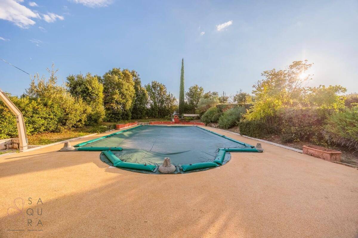 Großer Swimmingpool, abgedeckt mit einer grünen Plane, umgeben von einer gepflasterten Fläche und Bäumen.