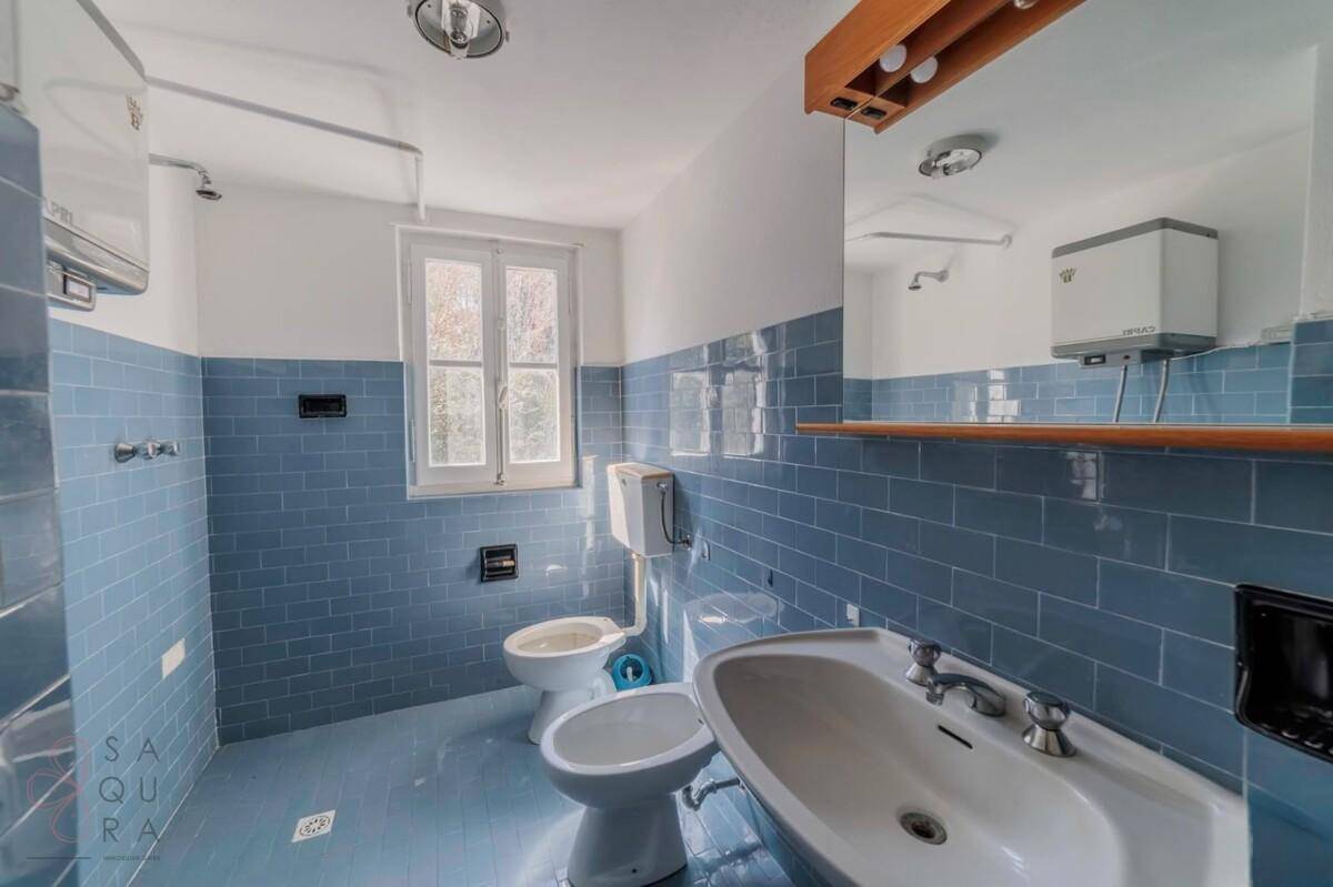 Badezimmer mit blauen Fliesen an den Wänden, einem Fenster, WC, Bidet und Waschbecken.