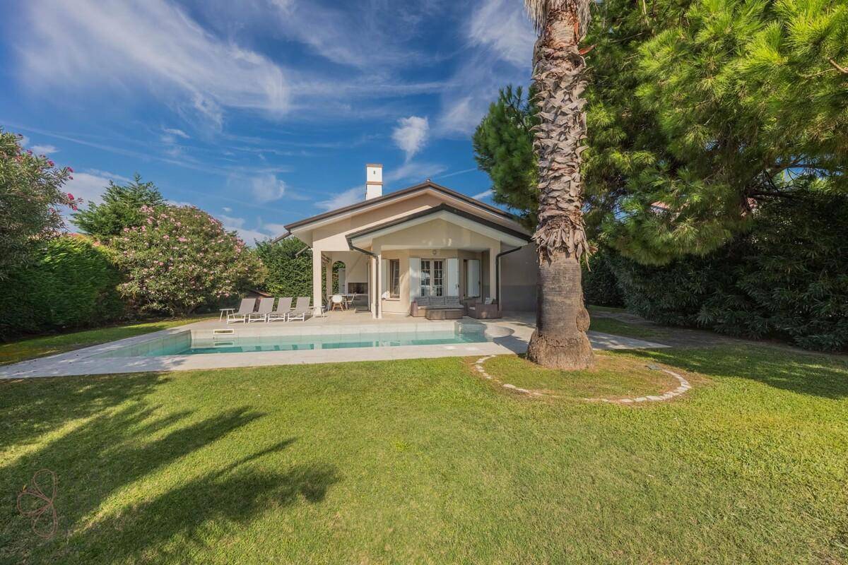Das Haus mit Pool und Garten, umgeben von hohen Bäumen und einer weitläufigen Rasenfläche unter strahlendem Himmel.