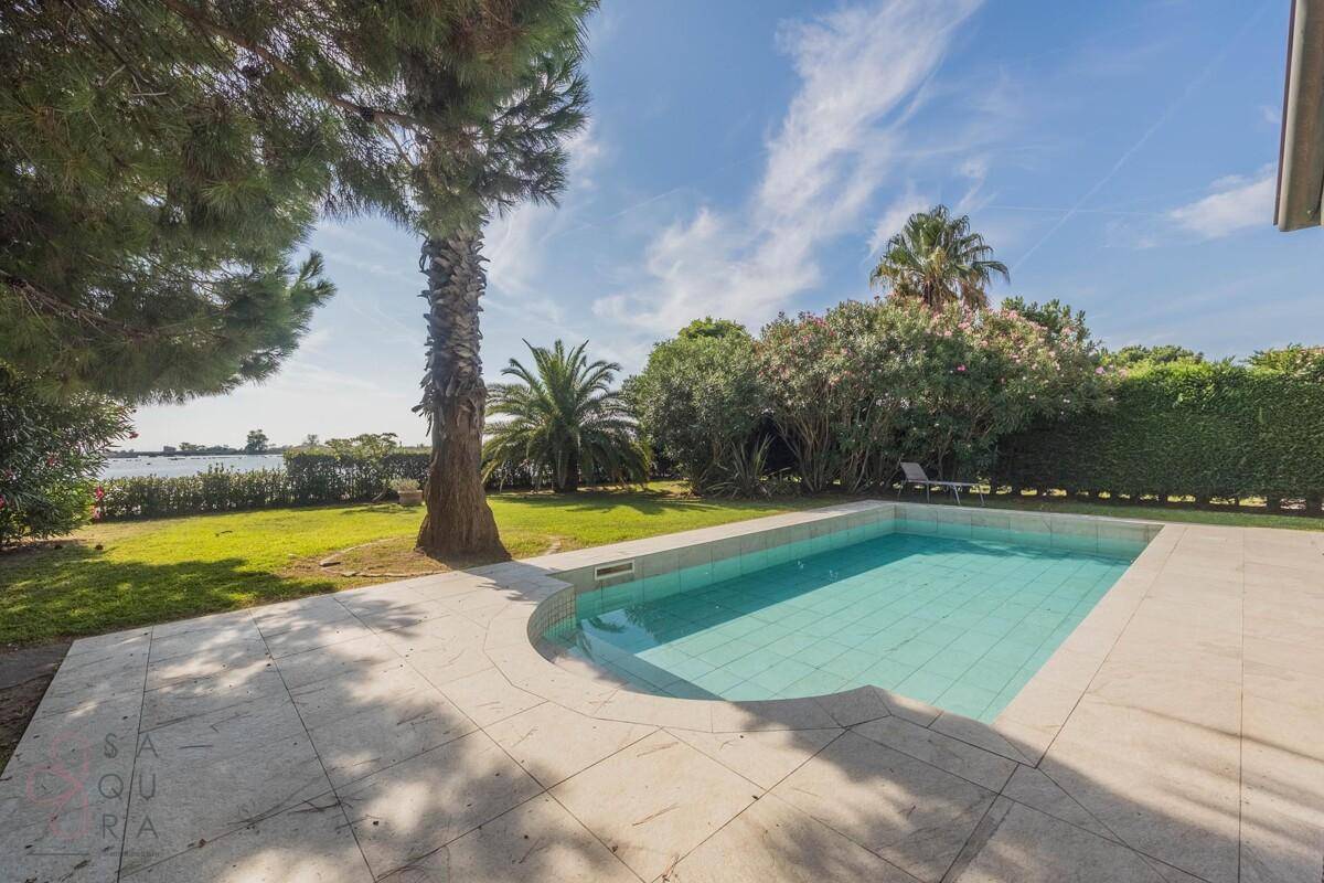 Poolbereich im Garten mit Blick auf das Wasser, umgeben von Palmen und grünen Hecken.