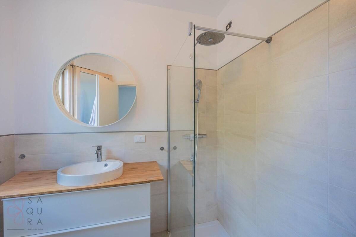 Modernes Badezimmer mit rundem Spiegel, Holzwaschtisch und ebenerdiger Dusche.
