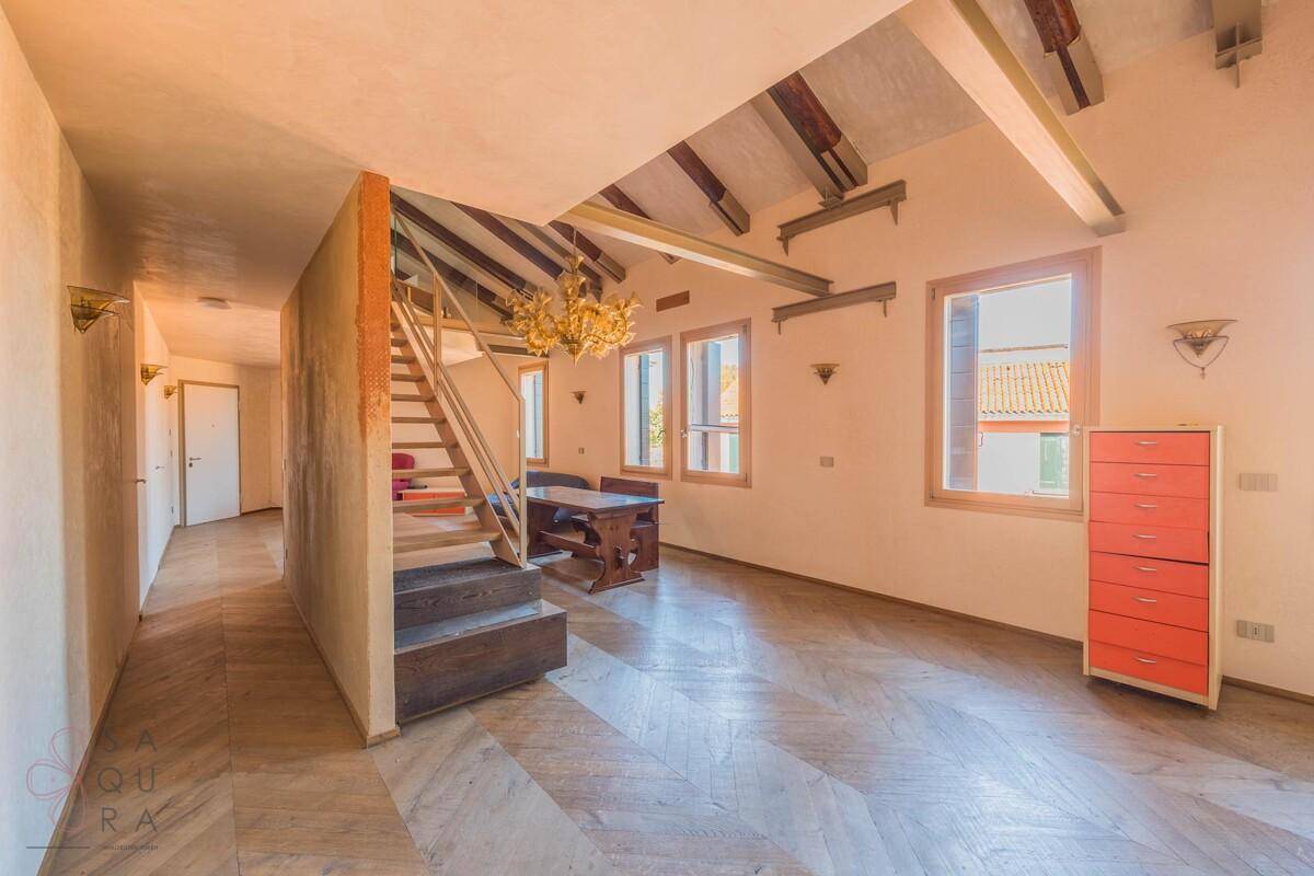 Heller Wohnbereich mit Fischgrätparkett, sichtbaren Holzbalken und einer markanten Treppe als zentrales Element.