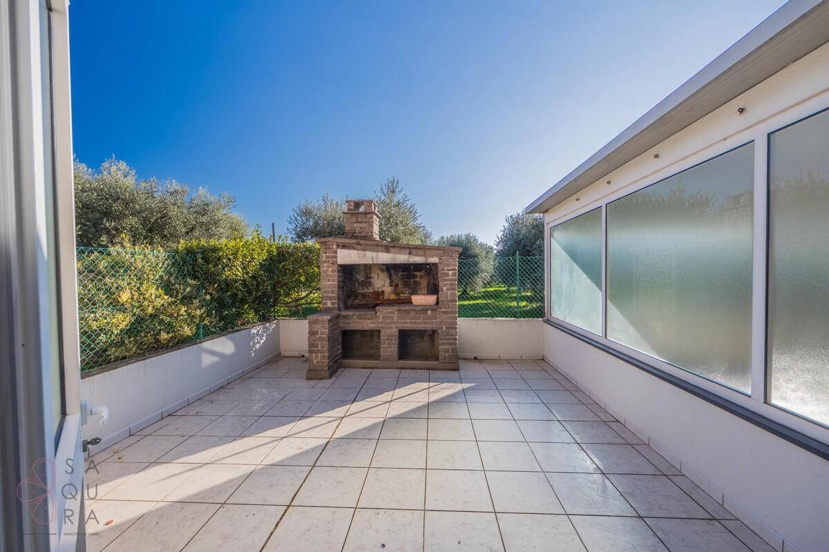 Sonniger Außenbereich mit gemauertem Grill und Fliesenboden, umgeben von grünen Hecken und Bäumen.