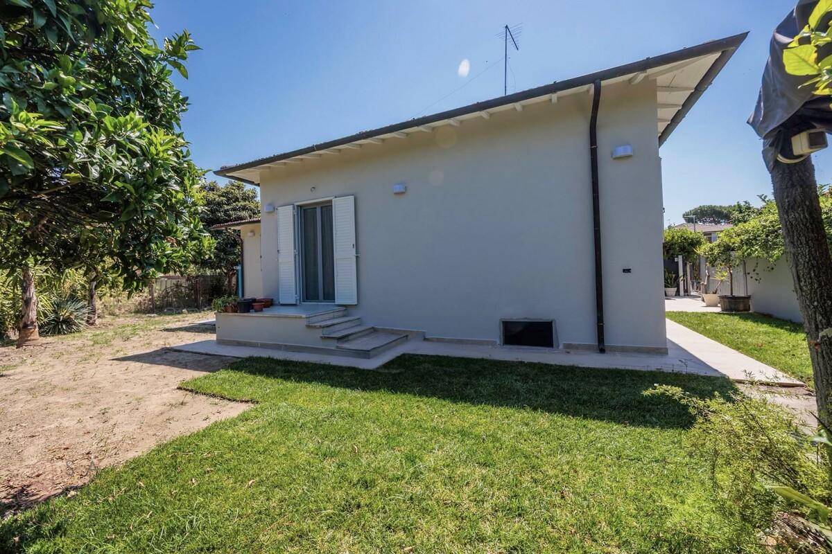 Rückseite des Hauses mit Rasenfläche, gepflastertem Bereich und Bäumen im Hintergrund.