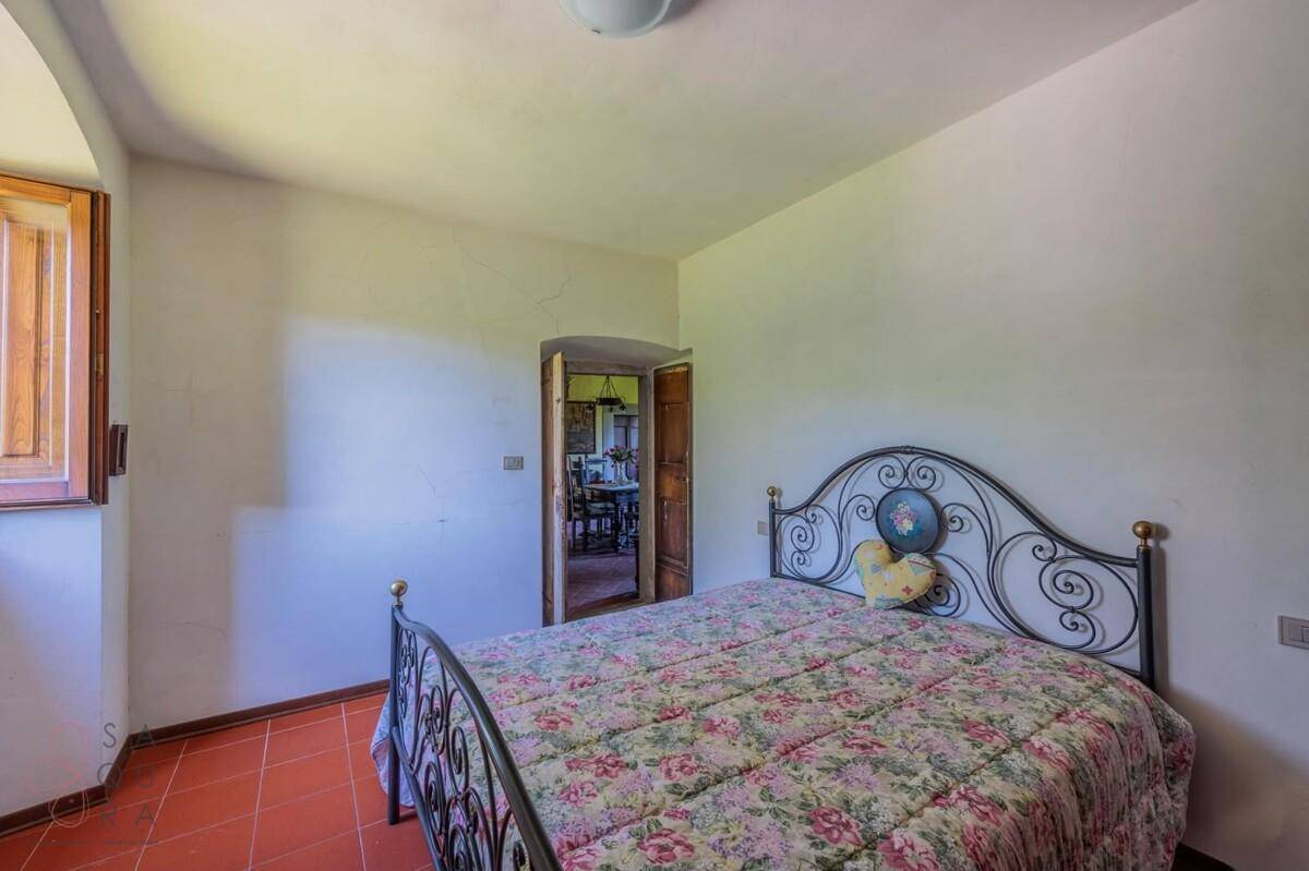 Schlafzimmer mit Terrakottaboden und schmiedeeisernem Bettgestell, das einen Blick in einen weiteren Raum bietet.