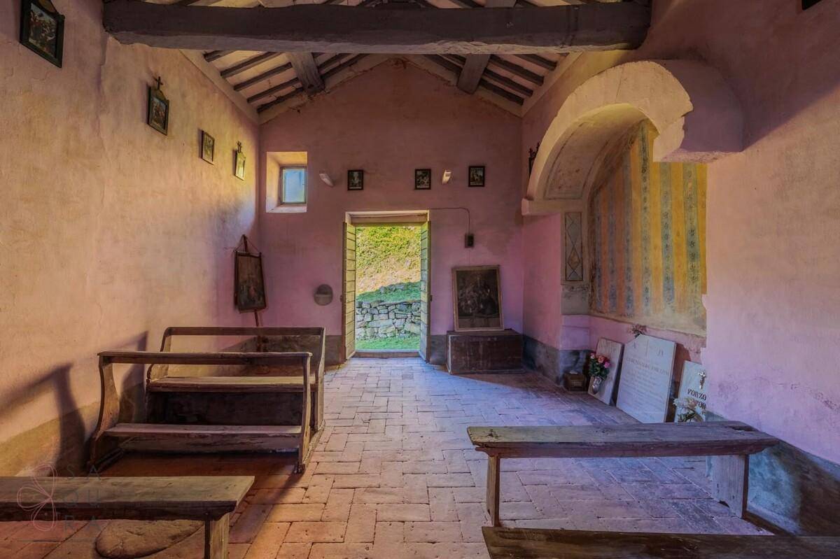 Innenansicht einer historischen Kapelle mit Holzbänken, rosa Wänden und Blick ins Grüne durch die Tür.