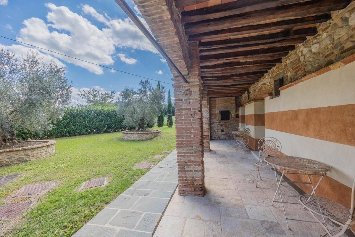 Überdachte Loggia mit rustikalen Möbeln und weitem Blick über die grüne Landschaft und Olivenbäume.