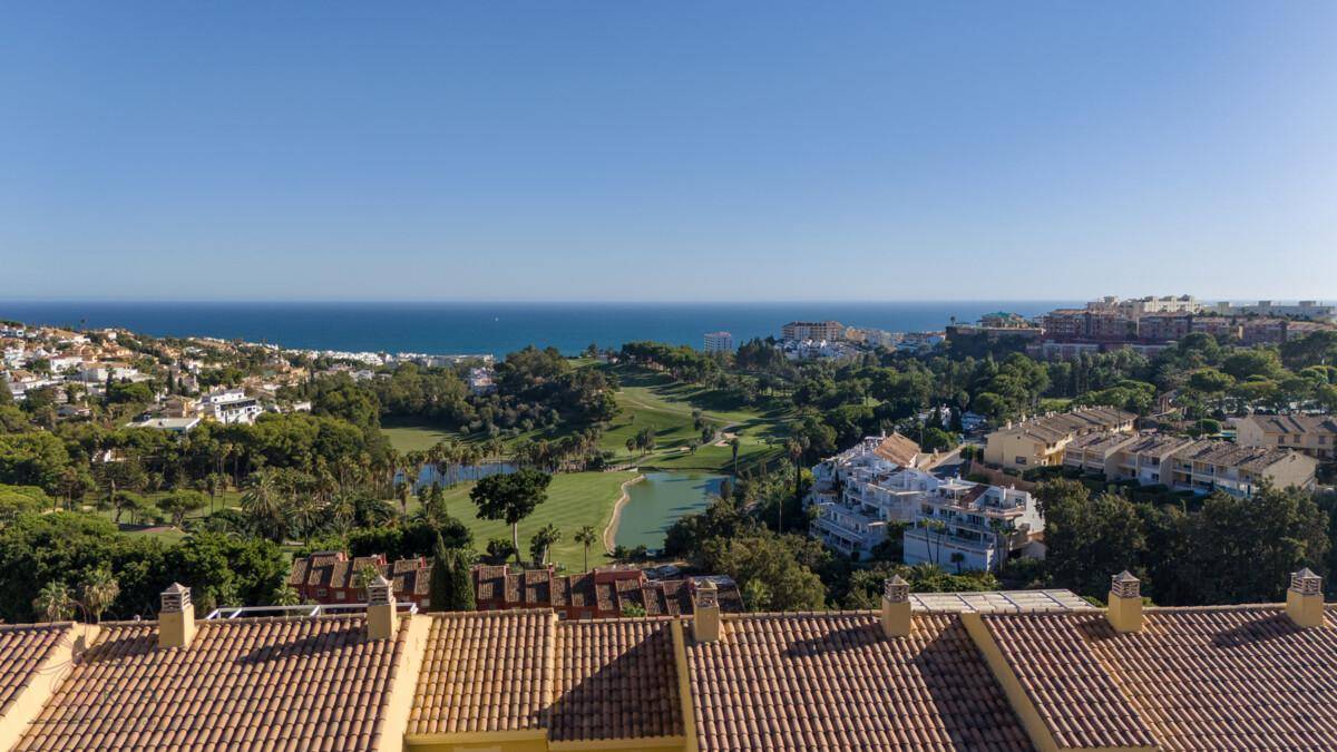 Weitblick über Ziegeldächer, einen grünen Golfplatz und das glitzernde Meer unter blauem Himmel.