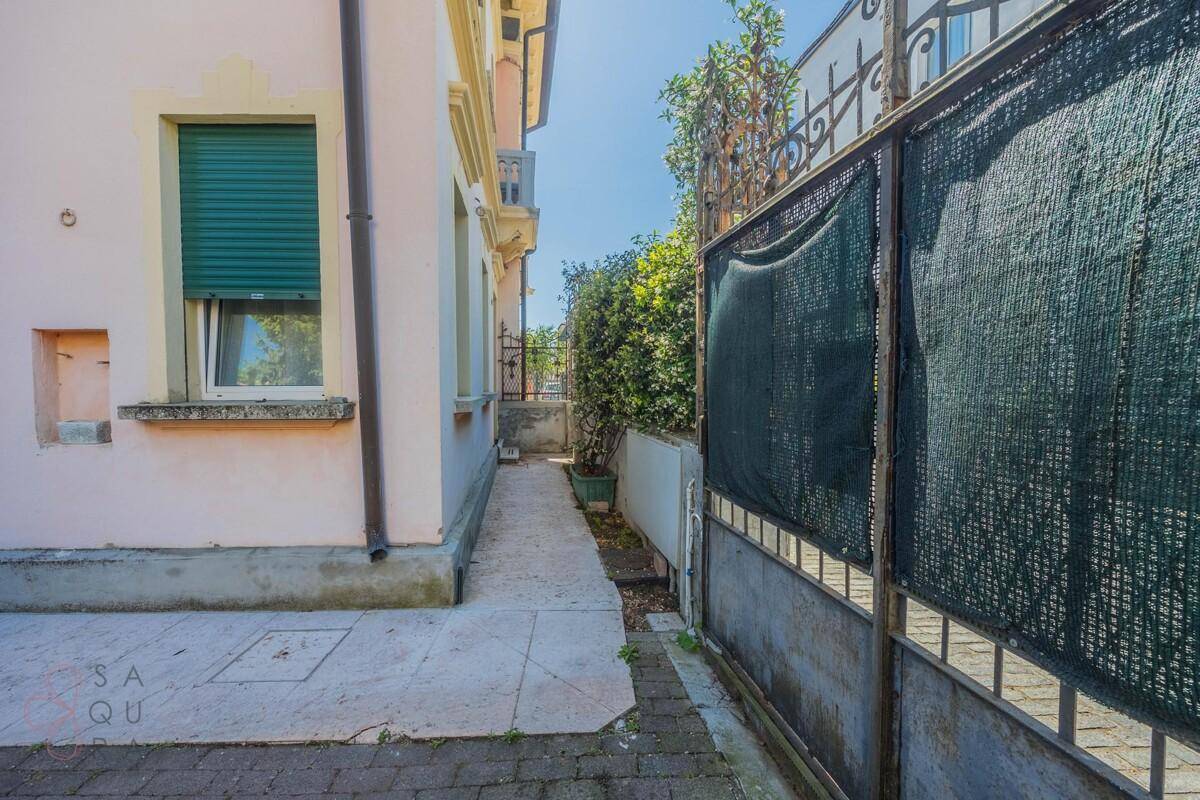 Schmaler Seitenweg entlang des Hauses mit einem grünen Sichtschutz am Zaun.