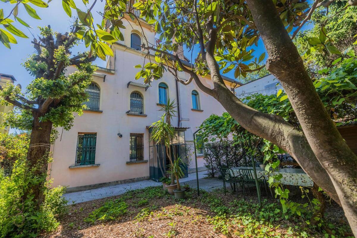 Grüner Gartenbereich hinter der Villa mit altem Baumbestand und einer schattigen Sitzgelegenheit.