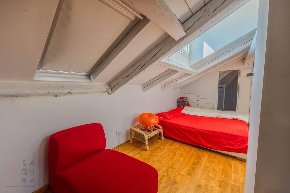 Schlafzimmer im Dachgeschoss mit Holzboden, rotem Bett und Dachfenstern für natürliches Licht.