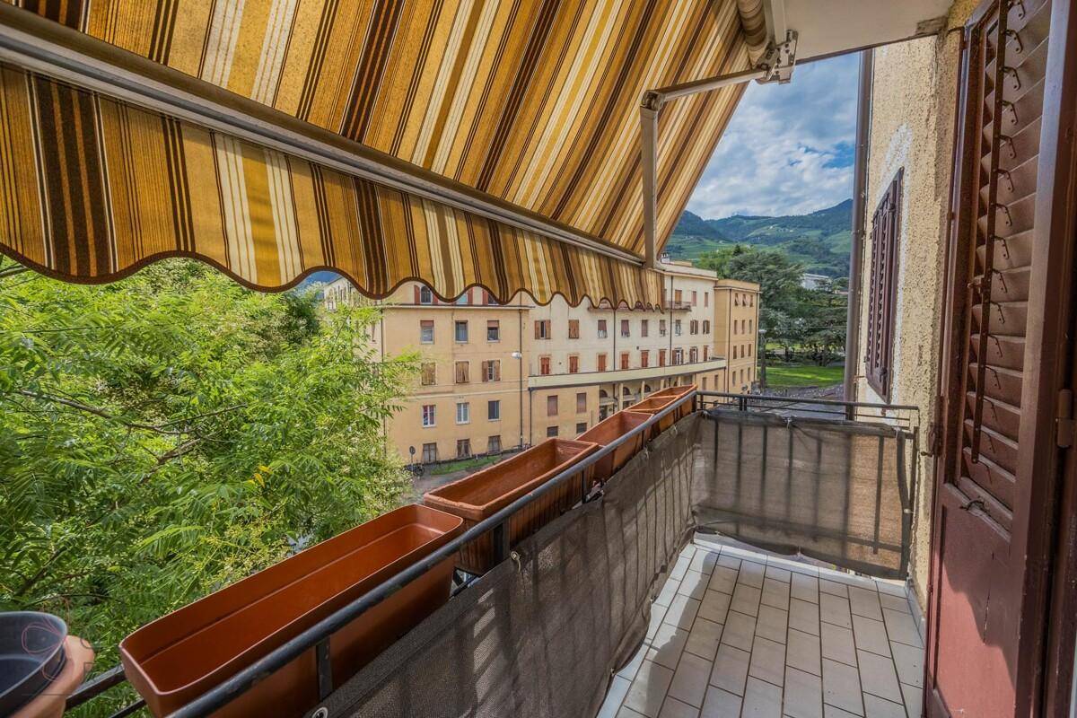Überdachter Balkon mit Markise, Fliesenboden und Blick auf umliegende Gebäude und Berge.