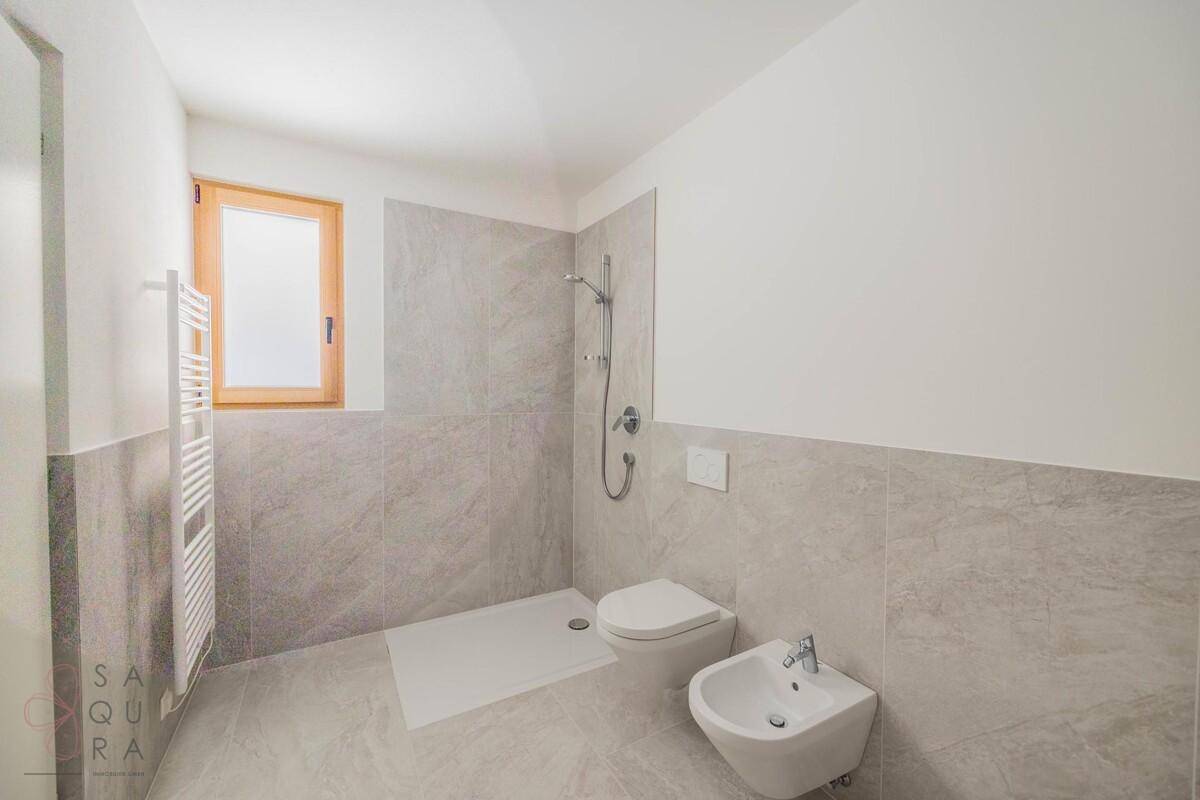 Modernes Badezimmer mit grauen Fliesen, ebenerdiger Dusche, WC, Bidet und Fenster.
