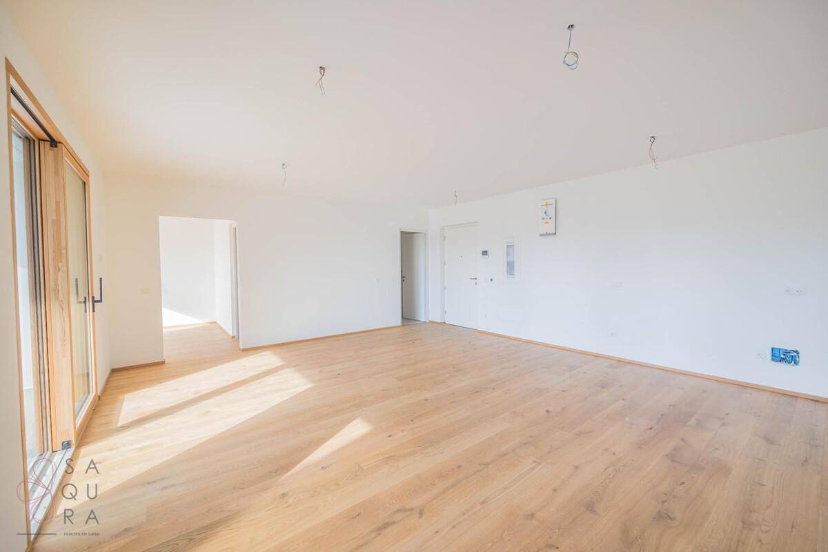Heller, unmöblierter Wohnraum mit Holzboden und großen Fenstern, die viel Tageslicht hereinlassen.