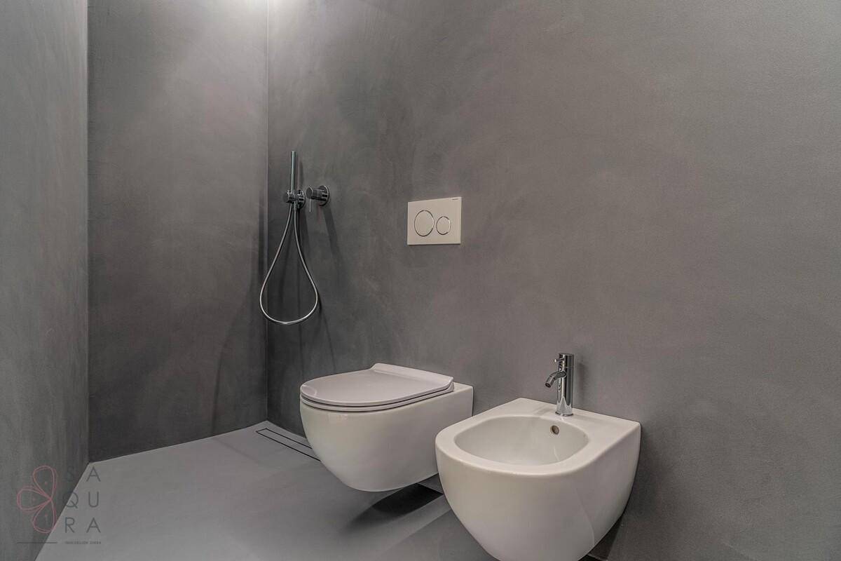 Stilvolles WC mit Bidet, wandhängender Toilette und Duschbereich in dunklem Grau.