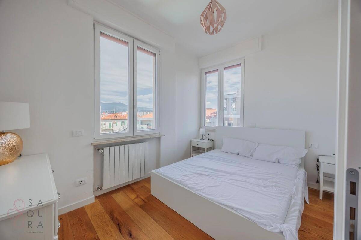 Schlafzimmer mit Doppelbett, Nachttisch und großen Fenstern, die viel Tageslicht hereinlassen.