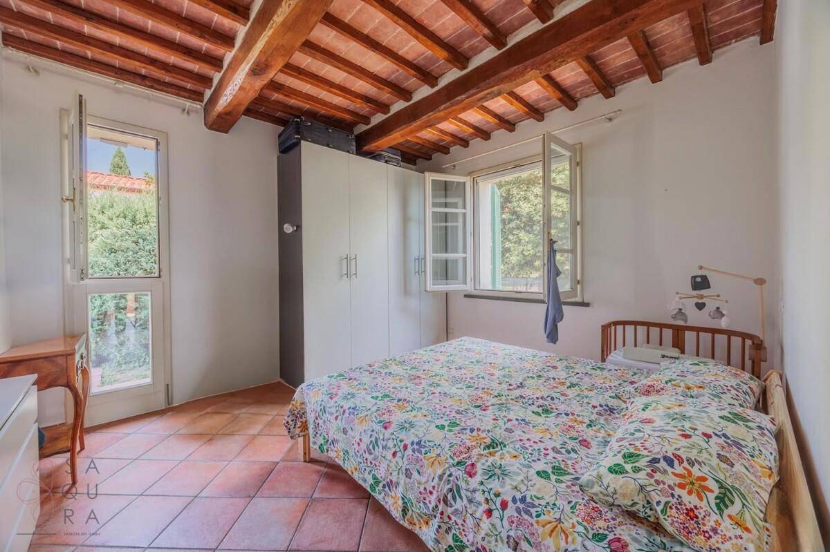 Helles Schlafzimmer mit Terrakottaboden, Holzbalkendecke und Blick auf die grüne Umgebung.