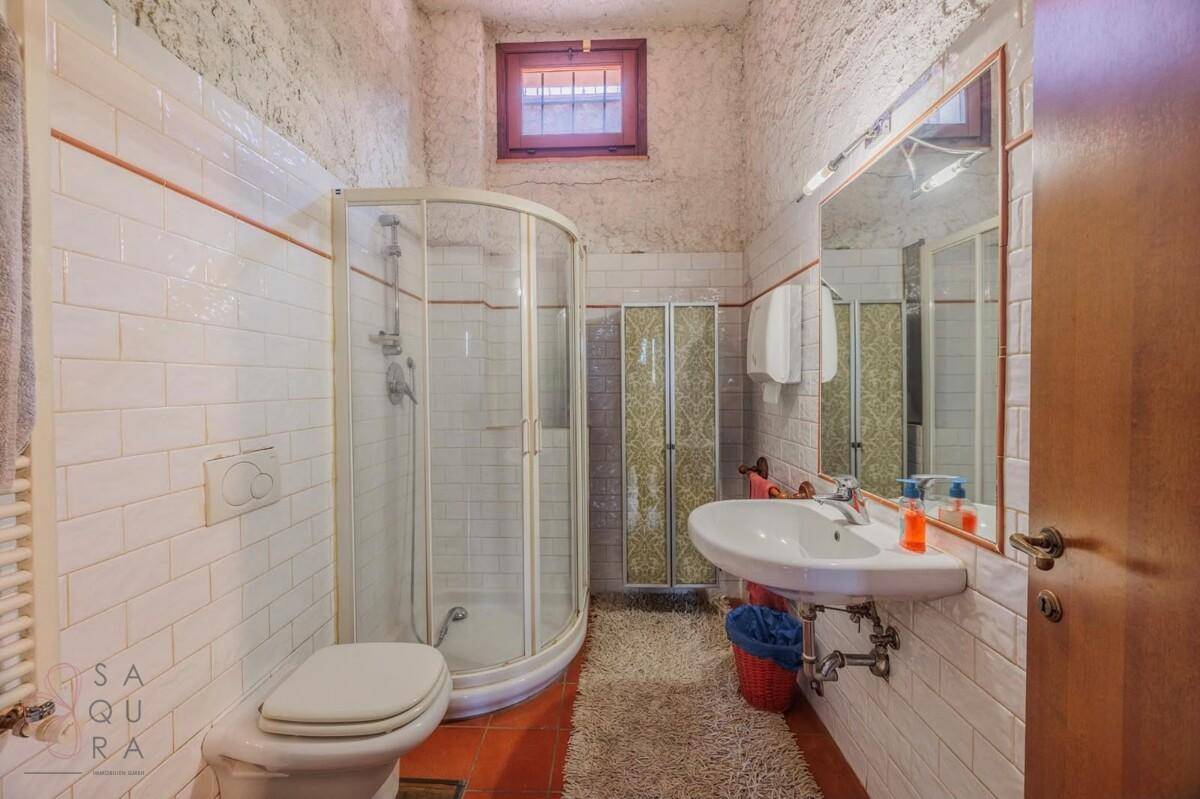 Badezimmer mit Eckdusche, WC und Einzelwaschbecken, ausgestattet mit weißen Fliesen und Terrakottaboden.
