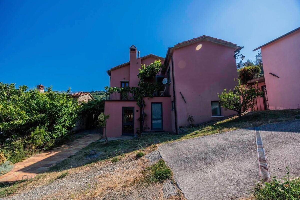 Rückseite des Landhauses mit rosa Fassade und Zugang zu einem kleinen Balkon, umgeben von Grün.