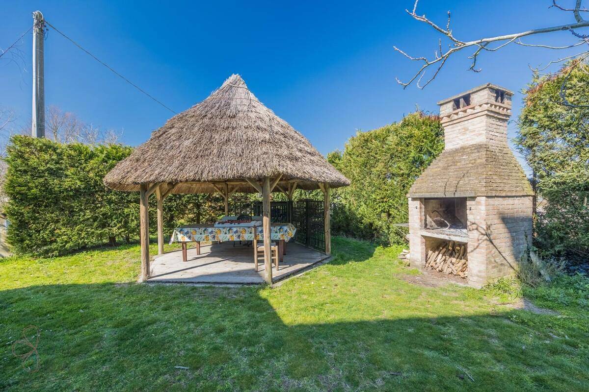Detailansicht des strohgedeckten Pavillons mit Esstisch und dem gemauerten Grill im weitläufigen Garten.