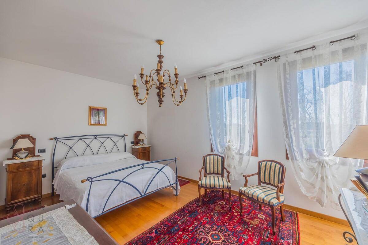 Schlafzimmer mit schmiedeeisernem Bett, Kronleuchter und zwei großen Fenstern mit Blick ins Grüne.