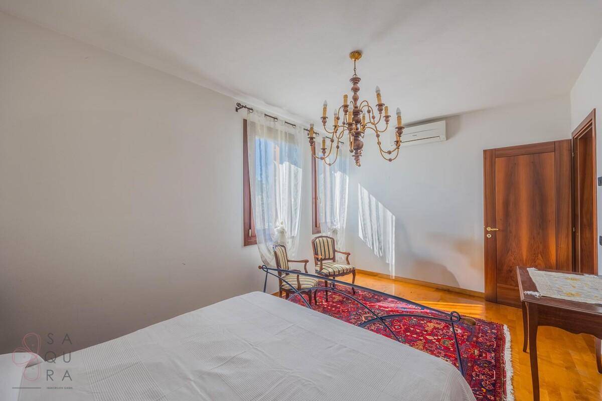 Helles Schlafzimmer mit Holzboden, Kronleuchter, Klimaanlage und Fenstern mit weißen Vorhängen.