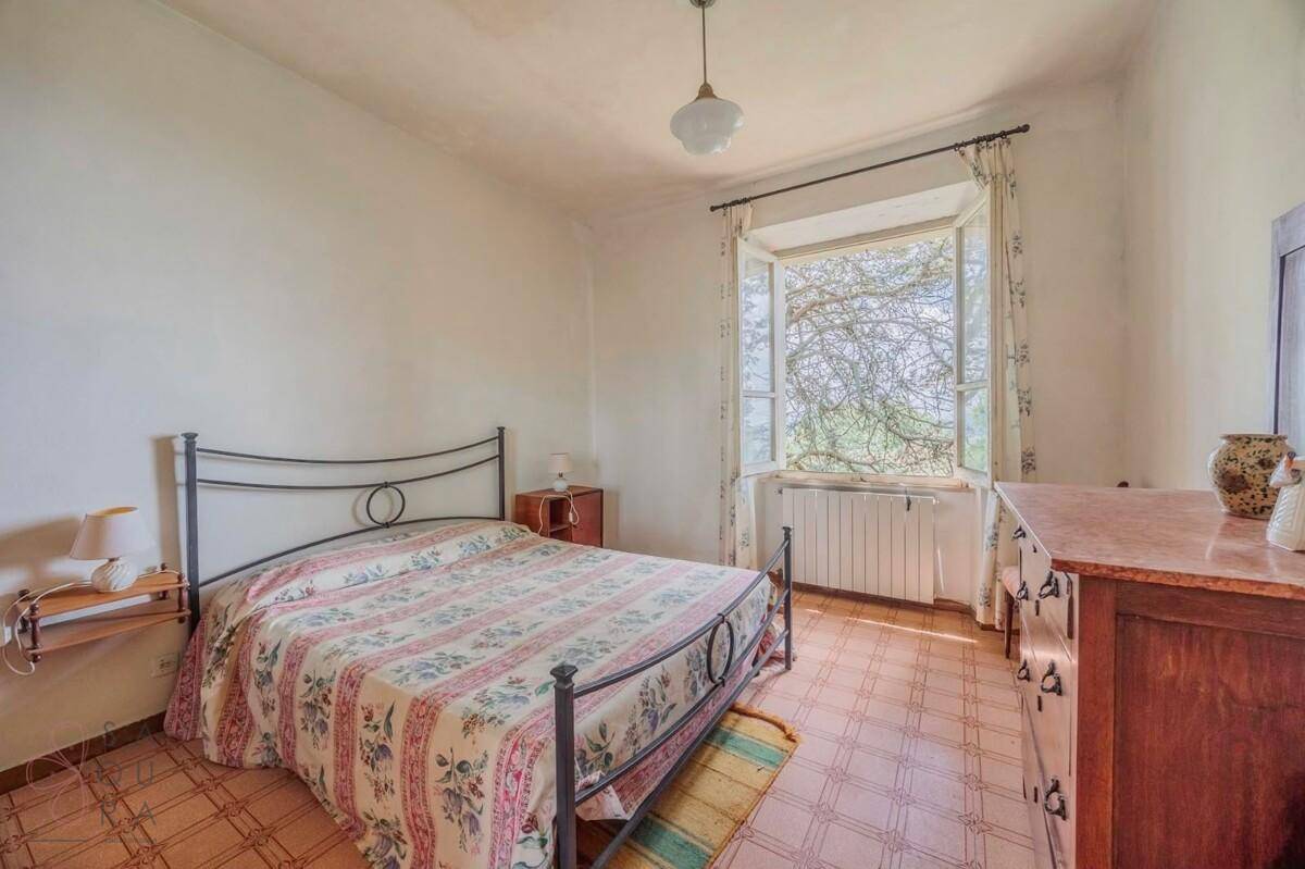 Schlafzimmer mit Metallbett, Holzkommode und Fenster mit Blick ins Grüne.