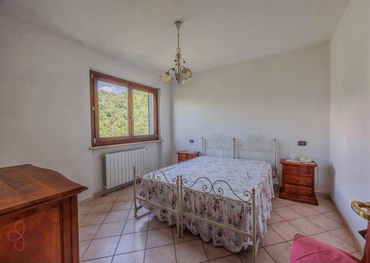 Schlafzimmer mit schmiedeeisernem Bett, Fliesenboden und Fenster mit Blick ins Grüne.