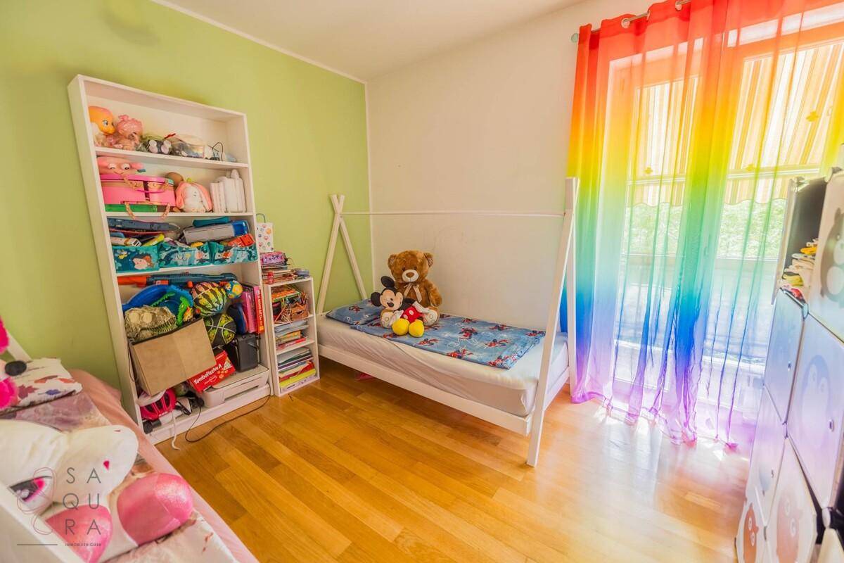 Helles Kinderzimmer mit zwei Betten, Bücherregal und farbenfrohen Regenbogenvorhängen.