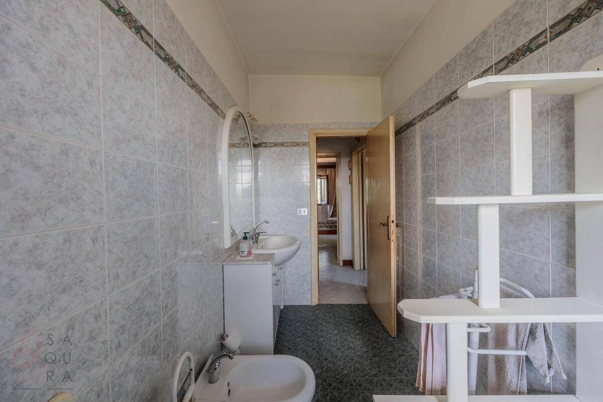Badezimmer mit grauen Fliesen, Einzelwaschbecken und einem Durchgang zu einem anderen Raum.