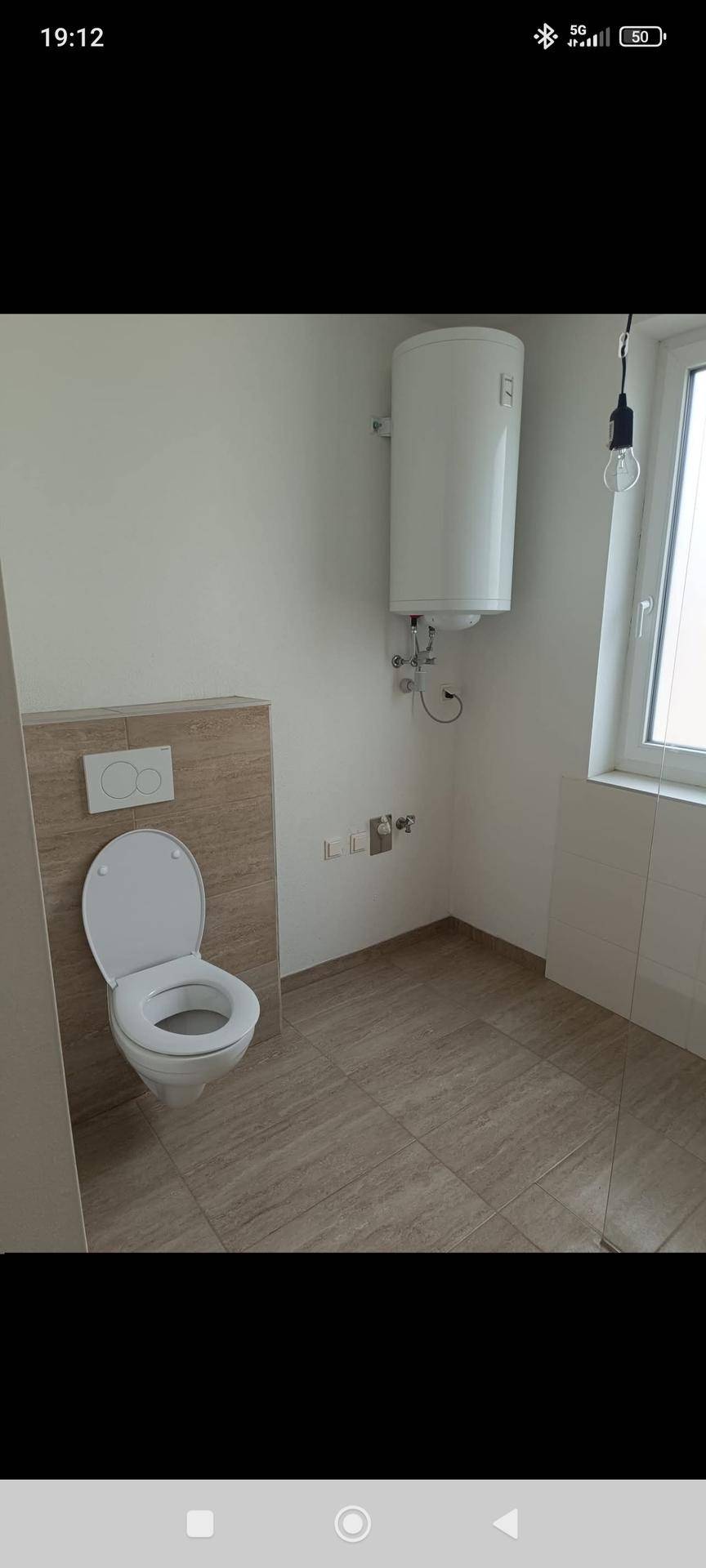 Helles WC mit wandhängender Toilette, Warmwasserboiler und Fenster, modern gefliest.