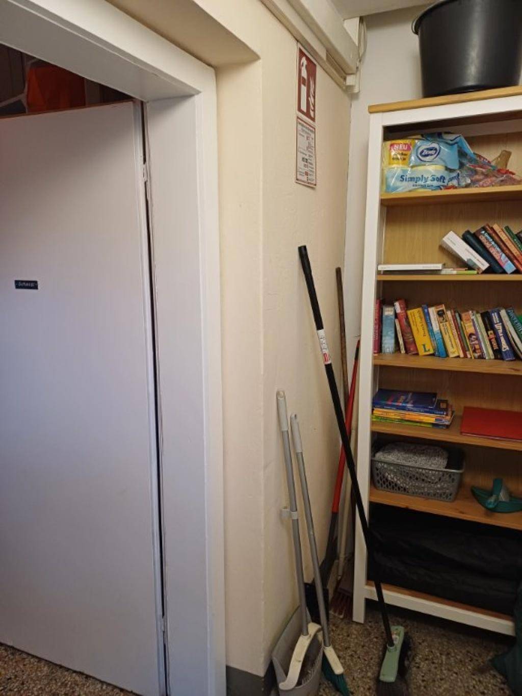 Flur mit Bücherregal und Reinigungsutensilien, angrenzend an einen weiteren Raum.