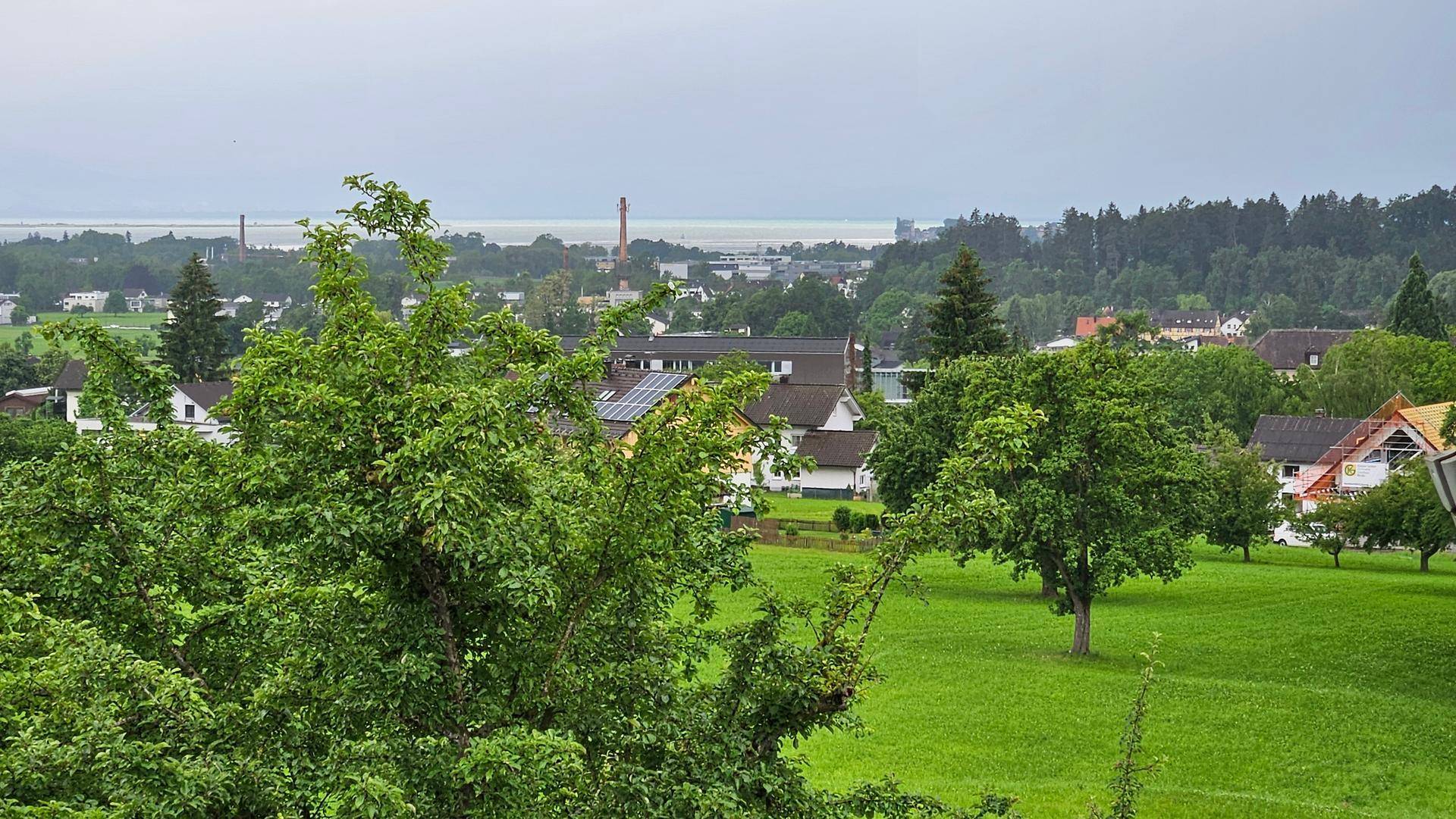 Weitläufiger Blick über grüne Landschaft, Häuser und einen See unter bewölktem Himmel.