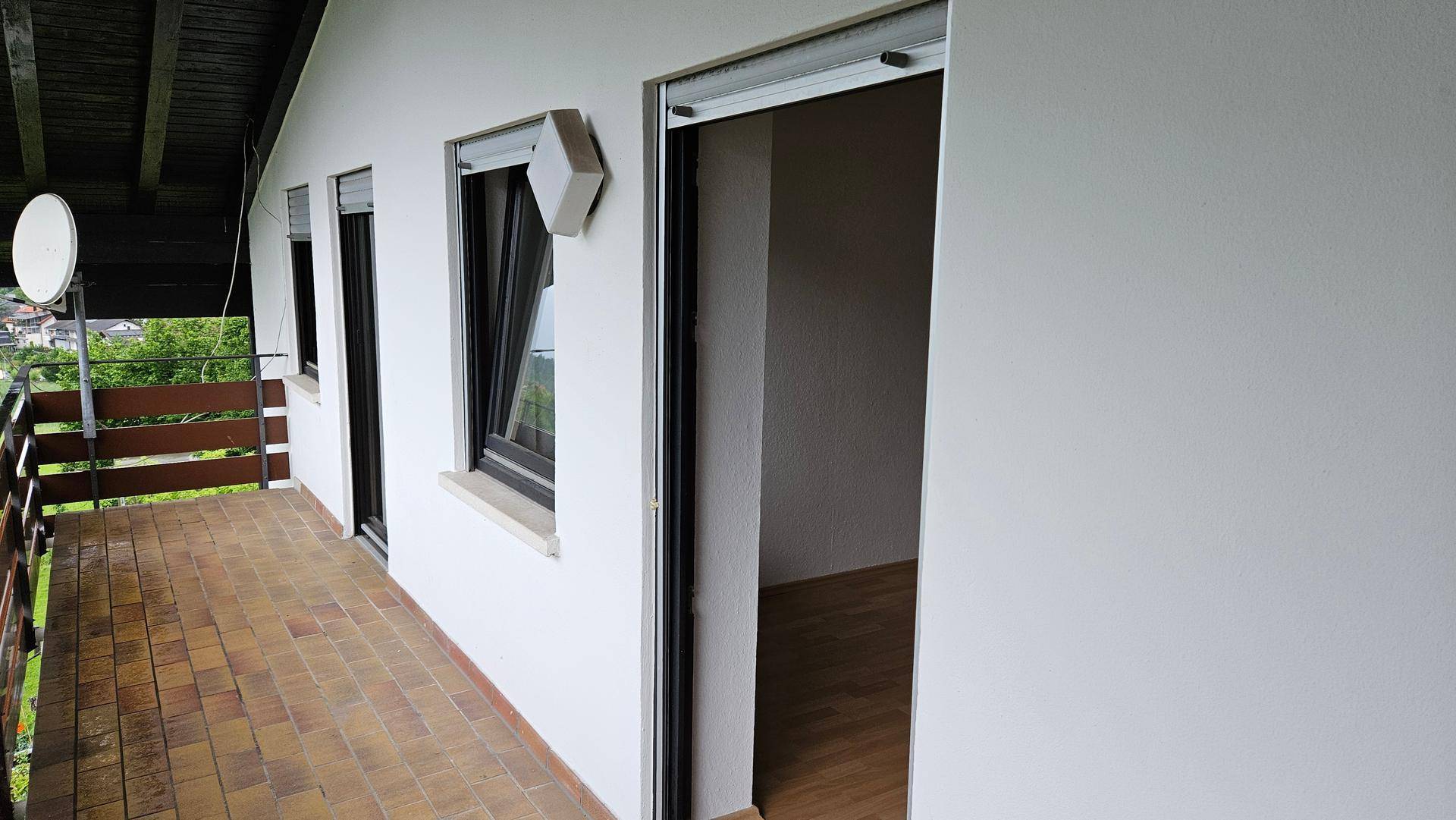 Langer Balkon mit Terrakottafliesen, Holzgeländer und Rollläden an den Fenstern.