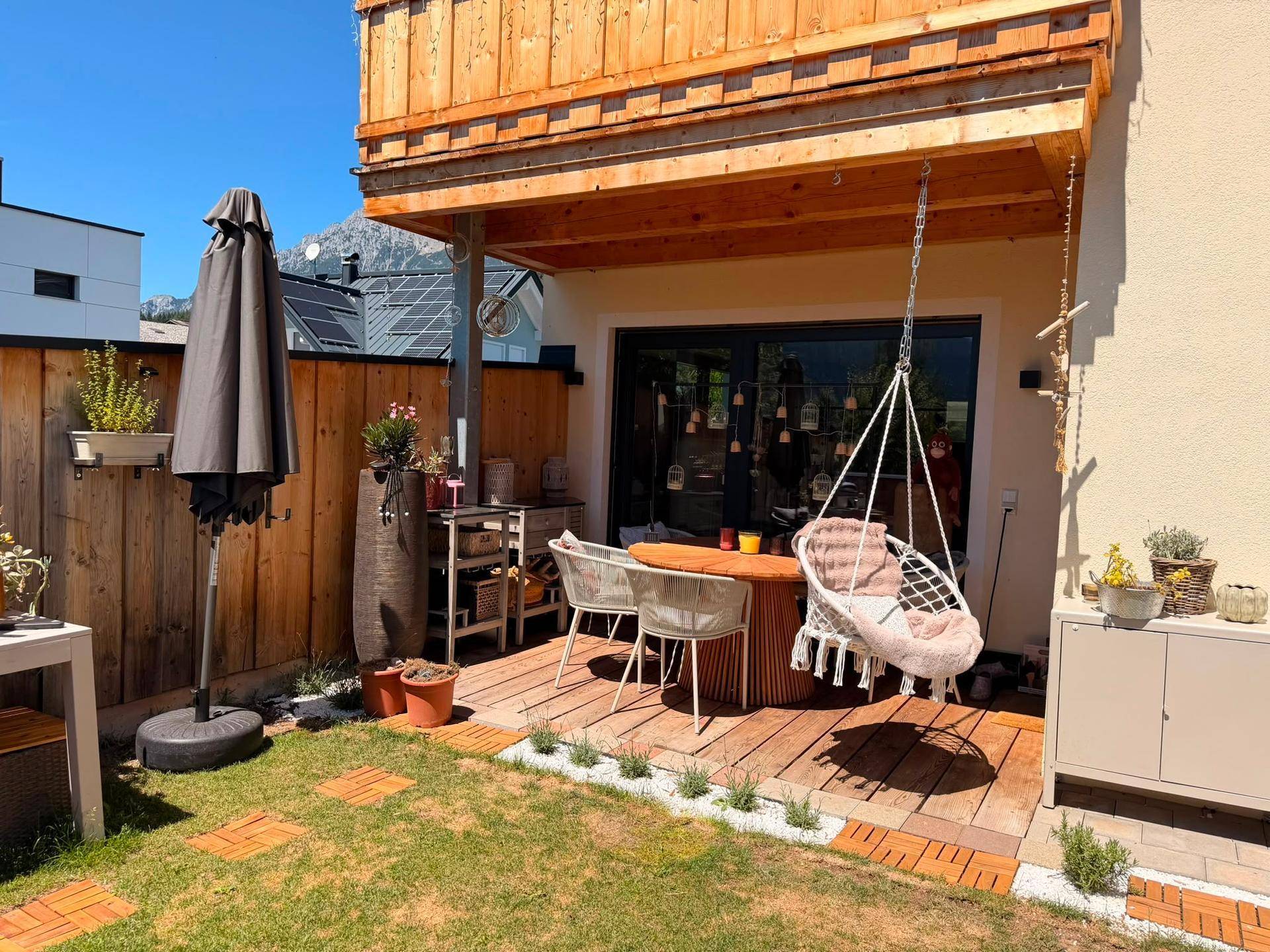 Sonnige Terrasse mit Holzdeck, Gartenmöbeln und Hängesessel, umgeben von Grün.
